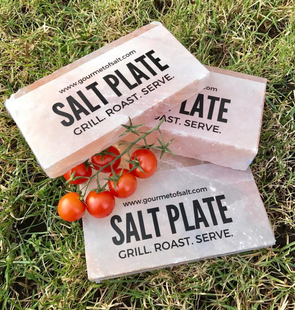 Three pink Himalayan salt plates with black text "SALT PLATE GRILL. ROAST. SERVE." stacked on green grass with a vine of red cherry tomatoes.