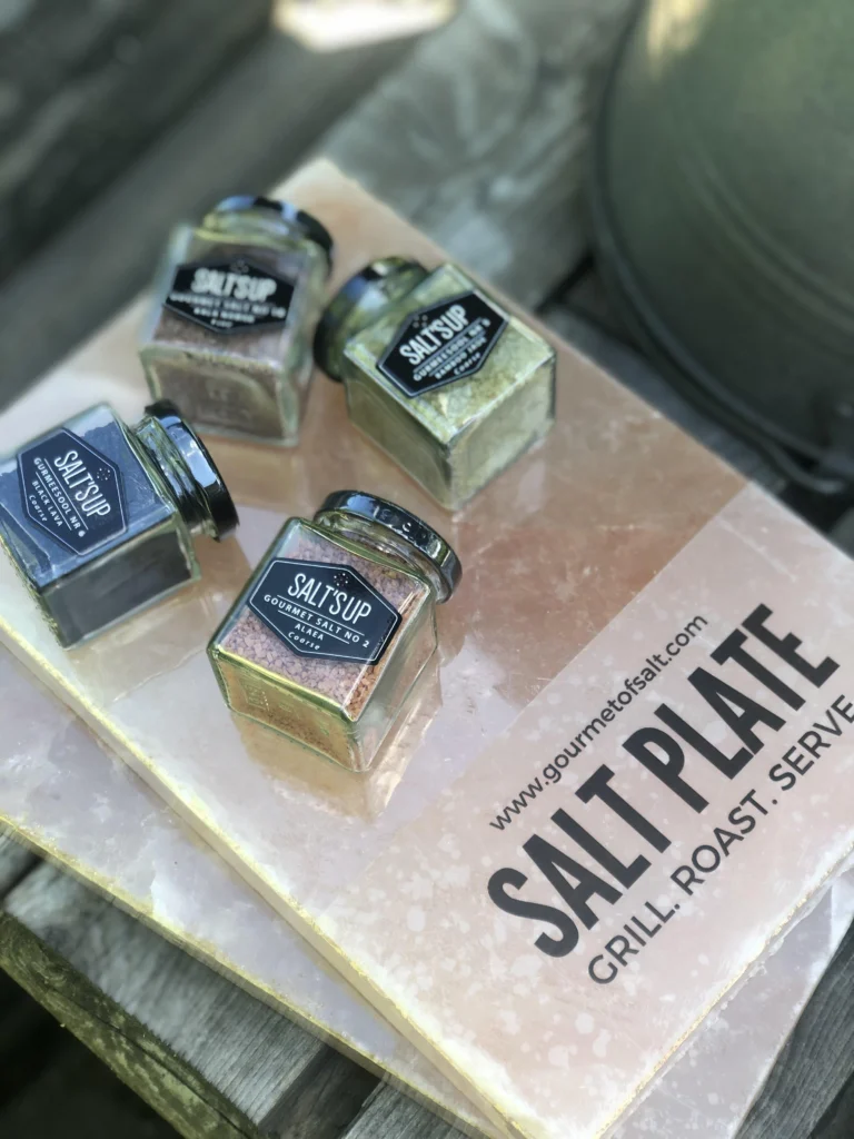 Close-up of a pink Himalayan salt plate with four small jars of 'Salt'sup' gourmet flavored salts resting on top, outdoors on a wooden surface.