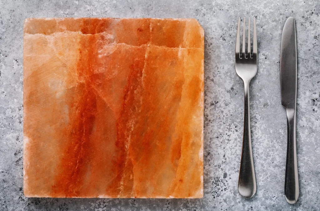 A square, rough-textured pink and orange Himalayan salt block next to a stainless steel fork and knife on a grey speckled counter.